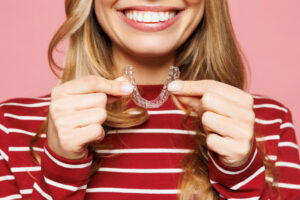 Close up cropped young smiling satisfied woman in red striped sweatshirt casual clothes hold in hand invisible transparent aligners, invisalign dental braces isolated on plain pink background studio